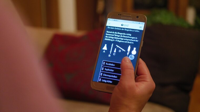 Symbolbild Actionbound Chemie: Eine Person hält ein Handy in der Hand und auf dem Bildschirm ist eine Quizfrage aus dem Bound zum Chemie-Studium zu sehen.