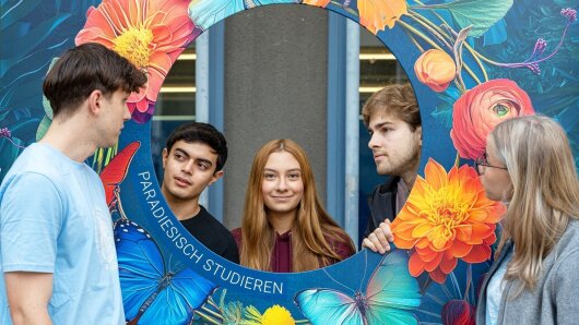 Fünf junge Menschen stehen auf dem Campus-Ernst-Abbe-Platz vor dem Schild "Paradiesisch studieren"