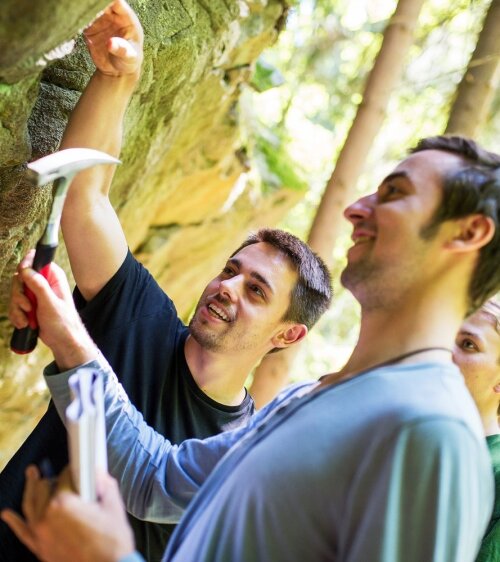 Zwei Studierende der Geowissenschaften der Universität Jena arbeiten im Gelände mit einem Geologenhammer.